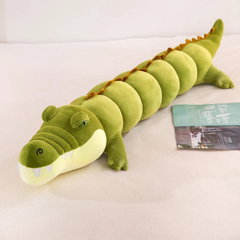 Green plushie crocodile body pillow on a white surface with a book next to it