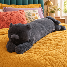 Moonlight Black Cat Body Pillow lying full length on mustard bedding in a colorful bedroom, showing its charcoal-black plush body, rounded head, small ribbed ears, gray muzzle, extended front paws, rear paws, and curled tail.