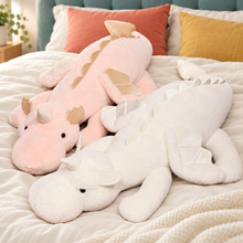 Pink and white Cuddle Soft giant dragon plushies lying side by side on ivory bedding, showing their soft wings, rounded snouts, and long floppy bodies.