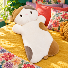 Calico lying cat plush pillow on mustard bedding with tan, gray and cream coloring.