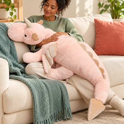 An adult lounges on an ivory sofa with the pink Cuddle Soft giant dragon plushie across her lap, showing its long body, wing, and tail.