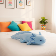 Blue axolotl plush toy on a bed with colorful pillows and a plant in the background.