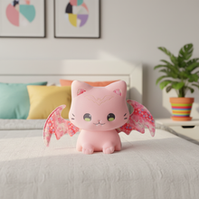 Pink cat with bat wings plush toy on a bed with colorful pillows and a plant in the background