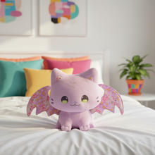 Purple cat plush toy with bat wings on a bed with colorful pillows and a plant in the background