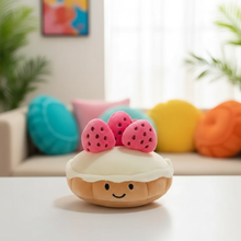 Plush toy resembling a dessert with strawberry on top in a room with colorful pillows.