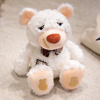 White teddy bear with a bow tie and band aid sitting on a carpeted floor.