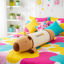 Capybara-shaped plush body pillow on a colorful bed with decorative pillows and plants in the background.