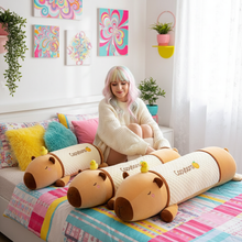 Woman sitting on a bed with large plush capybara toys and colorful pillows in a bright room.