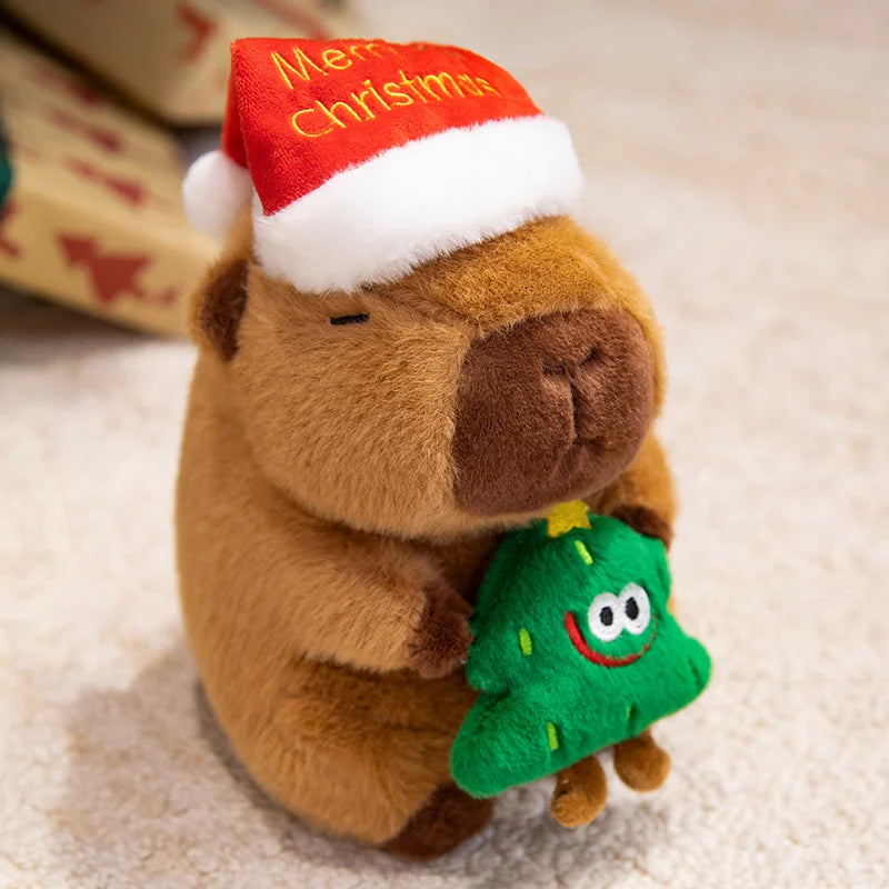 Plush capybara toy wearing a red 'Merry Christmas' hat holding a green tree toy on a soft surface.