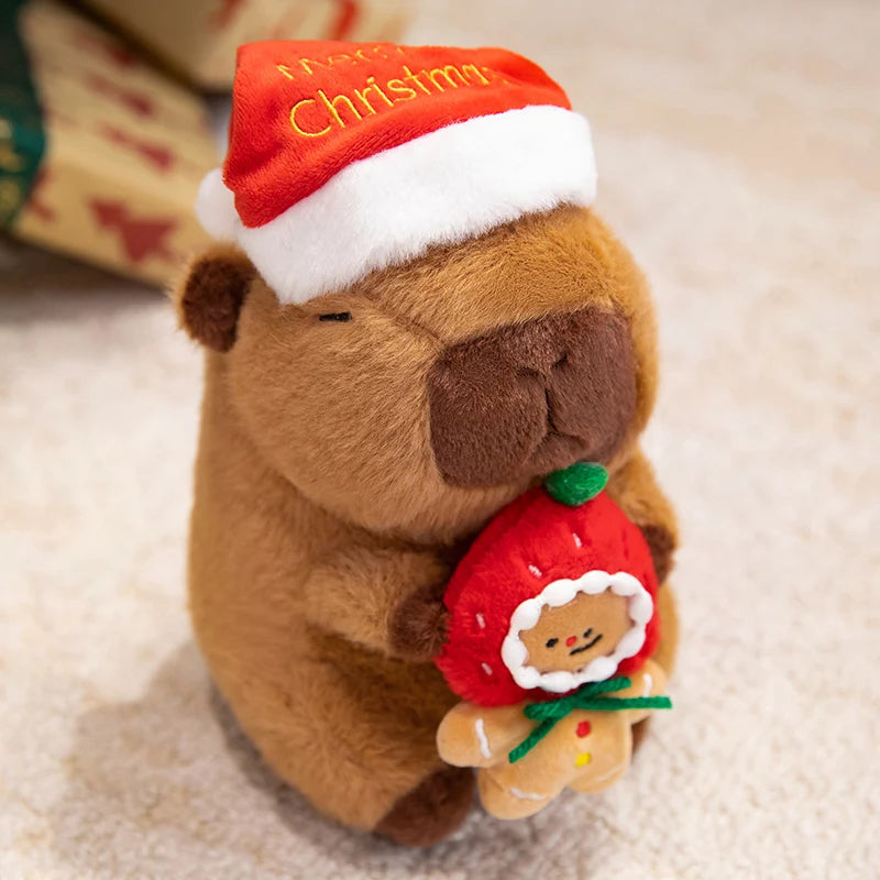 Plush toy capybara wearing a Santa hat holding a Christmas-themed ornament on a soft surface.