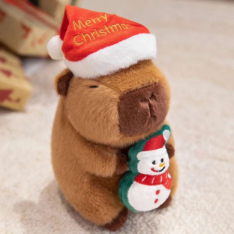 Plush capybara wearing a red 'Merry Christmas' hat and holding a snowman ornament on a light background.