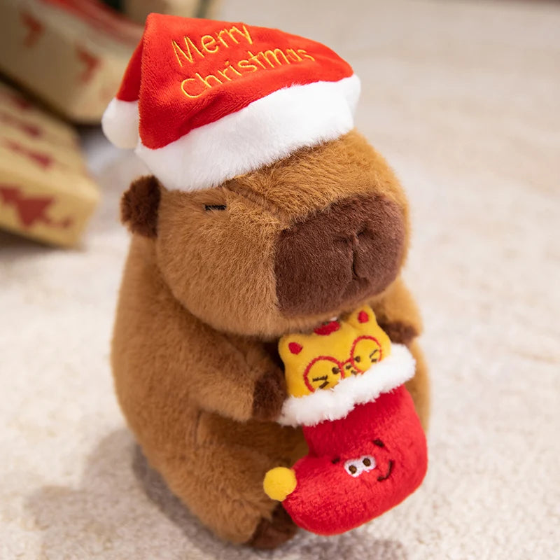 Plush toy capybara wearing a red 'Merry Christmas' hat and holding a red stocking with a small yellow cat inside.
