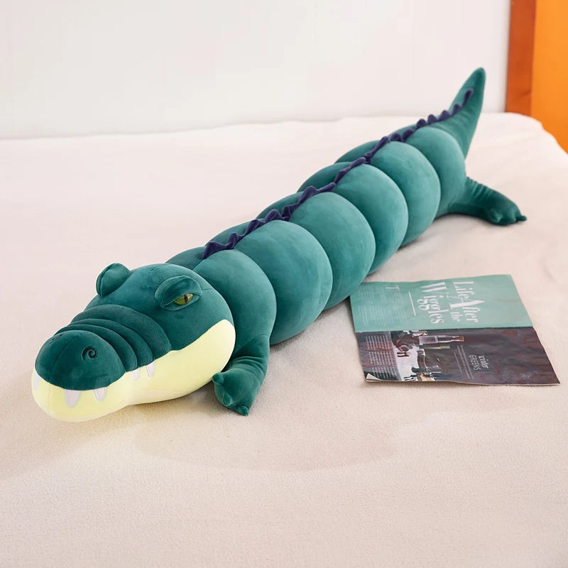 Green crocodile plushie body pillow on a bed next to a book