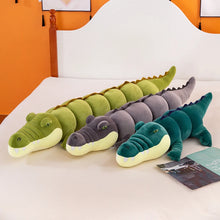 Three plushie crocodile toys in green, gray, and light green on a bed with an orange wall in the background.