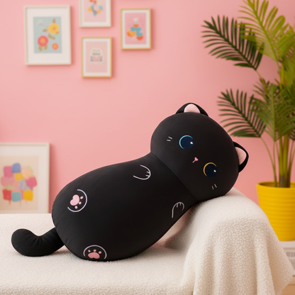 A black cat-shaped plush body pillow with colorful eyes and paw prints, resting on a white blanket against a pink wall with framed art and a potted plant.