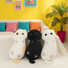 Three plush cat-shaped body pillows in white, black, and cream are arranged on a white sofa in a brightly lit living room with a yellow wall, colorful pillows, framed art, and a potted plant.