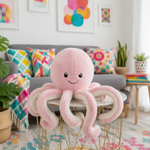 A pink plush octopus toy with a smiling face sits on a gold wire table in a bright, modern living room with a gray sofa, colorful pillows, and plants.