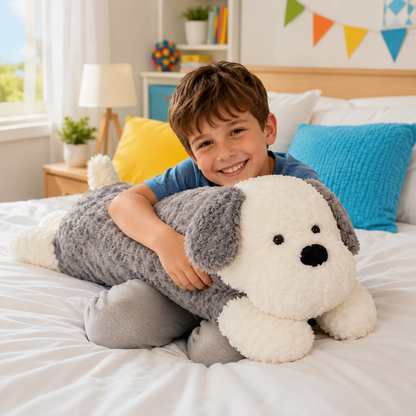A smiling young boy with brown hair sits on a white bed, hugging a large plush dog plushie body pillow. The dog is white with gray ears and paws, and has small black embroidered eyes and nose.