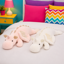Two plush dragon toys, one light pink and one white, lying side by side on a white bed with colorful pillows and a bright bedroom background.