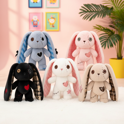 Four plush bunny toys in different colors and patterns are arranged on a white faux-fur surface against a soft pink room backdrop with framed art and a potted plant. The bunnies have long floppy ears, stitched facial details, and heart motifs on their chests.