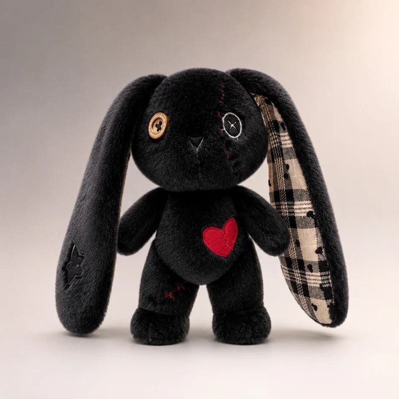A black plush bunny toy with long floppy ears, one ear showing a beige and black plaid lining, a red heart patch on the chest, and mismatched button eyes, photographed against a neutral beige studio background.