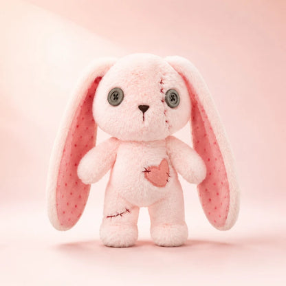 A pink plush bunny toy with long floppy ears, gray button eyes, and a small stitched heart on its chest, centered on a soft pink background.