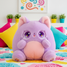 Purple plush cat with large eyes on a colorful blanket with pillows and plants in the background