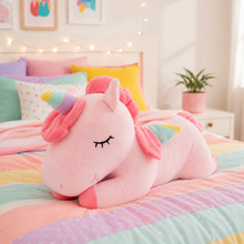 A pink plush unicorn pillow with a rainbow horn and pastel rainbow stripes, resting on a colorful pastel bedspread in a cozy bedroom setting.