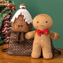 Gingerbread man plush toy with a small house plush toy on a wooden surface with a Christmas tree in the background.