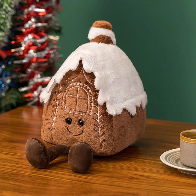 Plush gingerbread house toy with snow on a wooden table with a Christmas tree in the background.