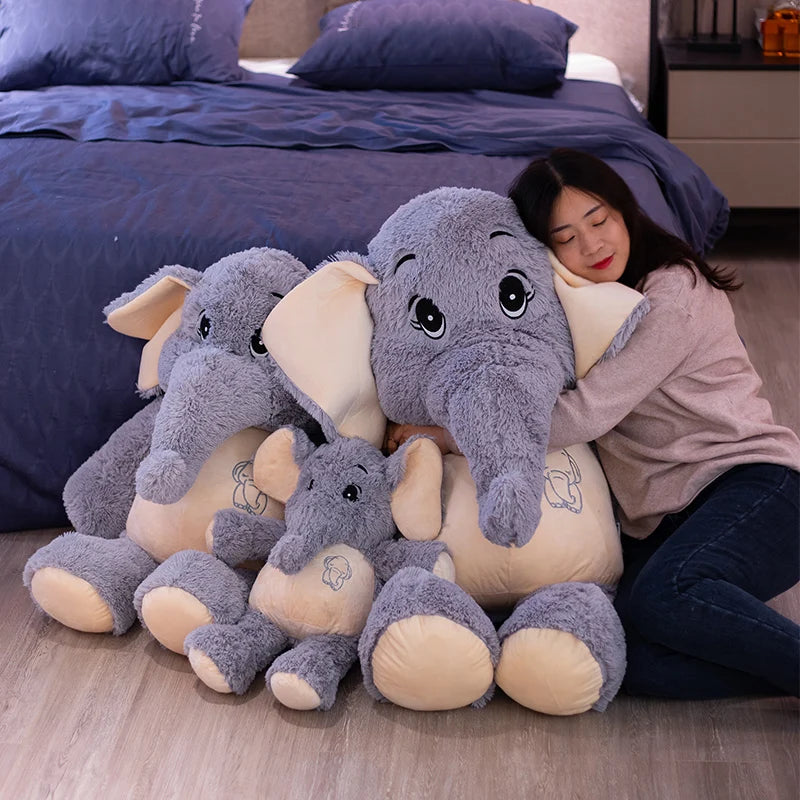 Woman hugging a large plush elephant toy with smaller ones on a wooden floor.