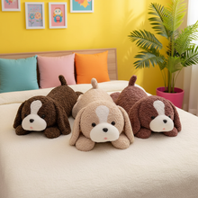 Three plush puppy toys in brown and beige are arranged on a bed in a brightly lit bedroom with a yellow wall, colorful pillows, framed art, and a potted plant.