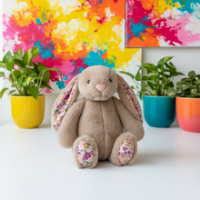 Plush bunny with floral ears and feet in front of colorful abstract art and potted plants.
