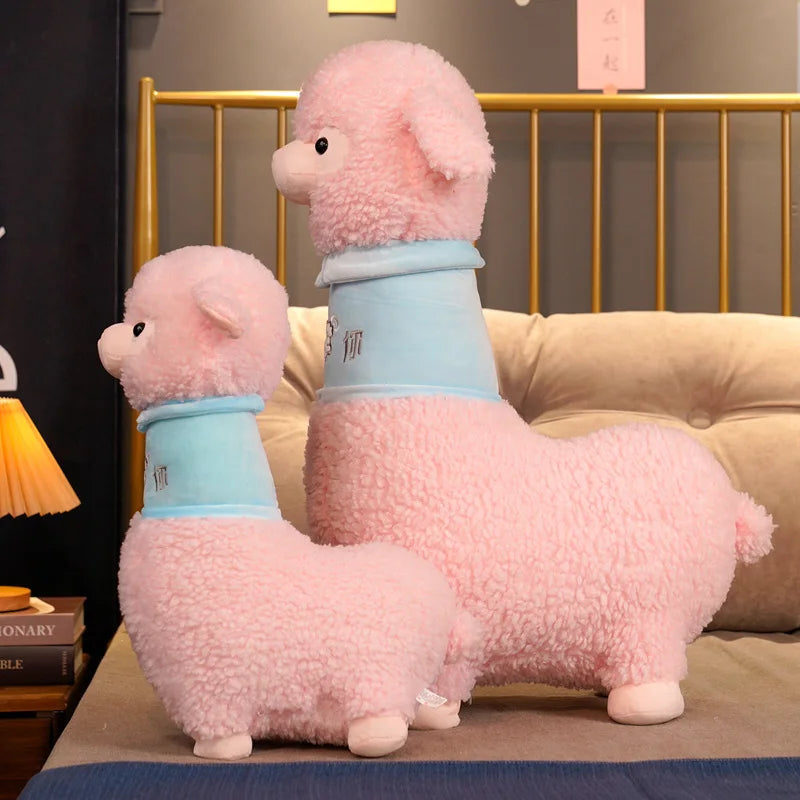 Two pink plush llamas with blue scarves on a bed.