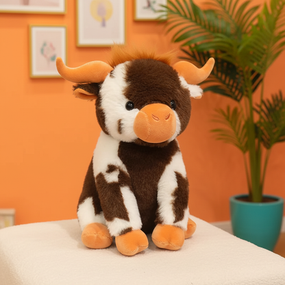 A brown and white cow plush toy with orange horns and a peach-colored snout, sitting on a white surface against an orange wall with framed pictures and a potted plant in the background.