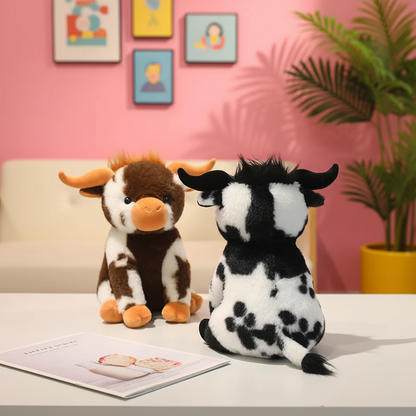 Two plush cow toys on a white table in a styled room setting with a pink wall, framed art, and a potted plant. One cow is brown and white facing forward, and the other is black and white facing away.