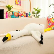 Large flamingo plush toy on a couch with colorful pillows and plants in the background