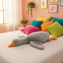 Gray plush goose toy on a bed with colorful pillows in a bright room.