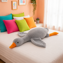A large plush gray duck toy with an orange beak and orange webbed feet lies on a white bed in a brightly lit bedroom. Colorful pillows in teal, pink, lime green, and orange are arranged behind it against a peach wall.