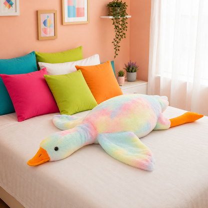 A pastel rainbow plush goose toy with an orange beak and feet, laid across a bed with colorful pillows in a bright bedroom setting.