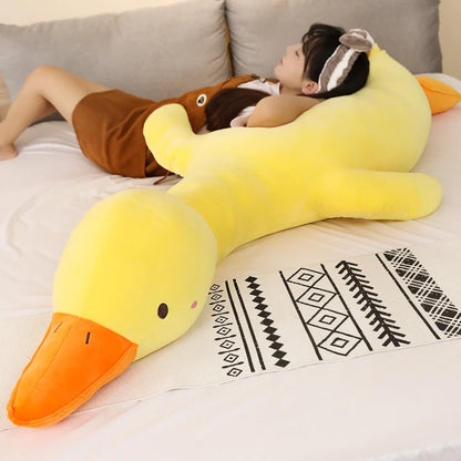 Large yellow duck-shaped pillow on a bed with a woman lying behind it.