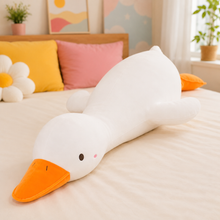A long white duck-shaped plush body pillow with an orange beak and orange tail, lying on a bed in a softly decorated bedroom.