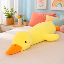 A large yellow duck-shaped plush body pillow with an orange beak lies stretched across a bed in a bright, pastel bedroom. The room includes colorful pillows, potted plants, and framed wall art.