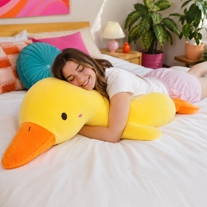 A woman smiling while lying on a bed and hugging a large yellow duck plush body pillow with an orange beak.