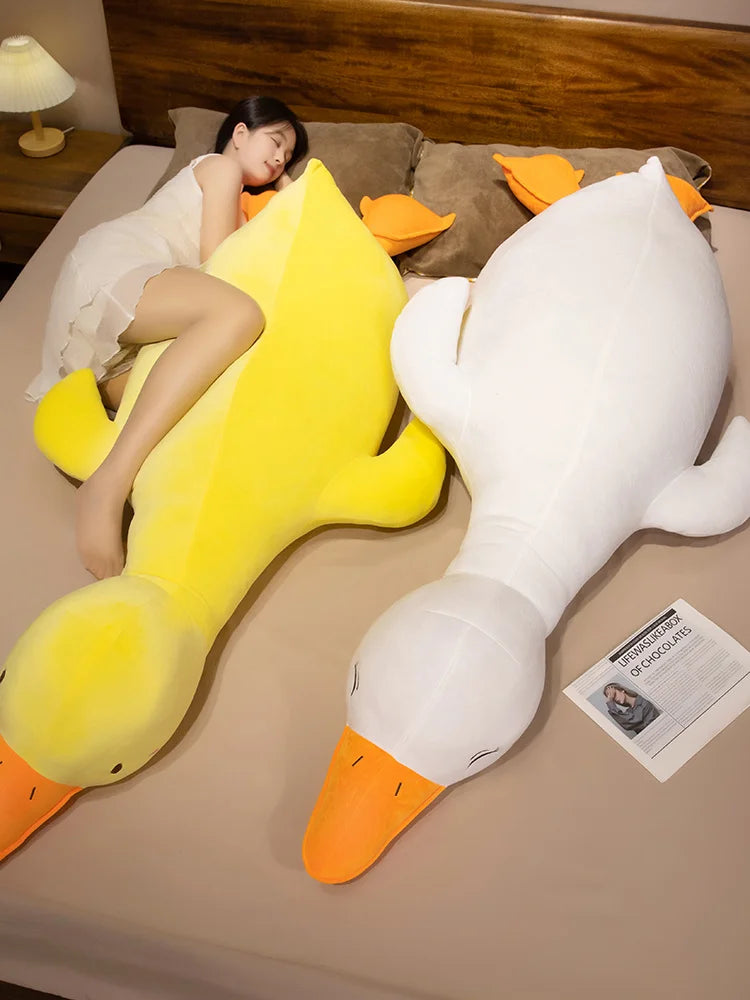 Large yellow and white goose plushie body pillows on a bed with a person lying next to them.