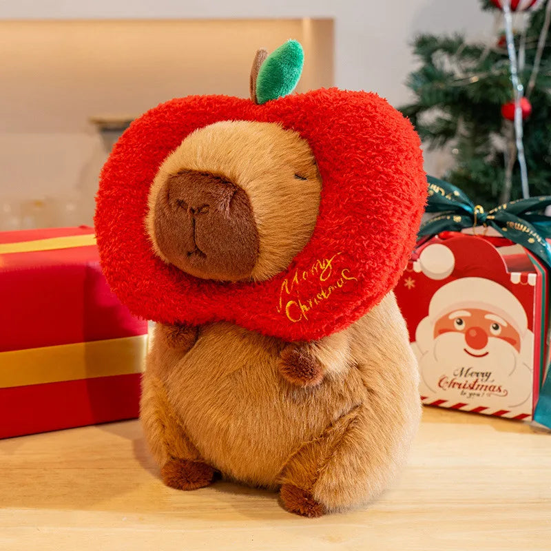 Plush toy capybara wearing a red 'Merry Christmas' collar with Christmas presents and decorations in the background.