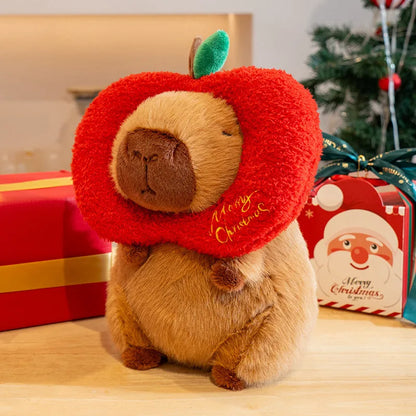 Plush toy capybara wearing a red 'Merry Christmas' collar with Christmas presents and decorations in the background.