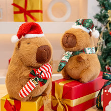 Two capybara plushies in festive attire on Christmas presents with a decorated tree in the background.