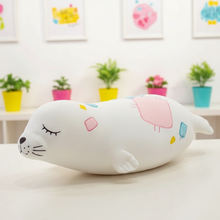 White plush toy with colorful accents on a light surface, with a blurred background of plants and framed pictures.