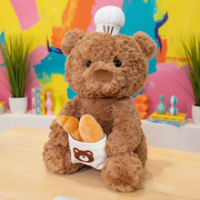Plush teddy bear with a chef's hat on its head and a small bag with a bear emblem, set against a colorful abstract background.